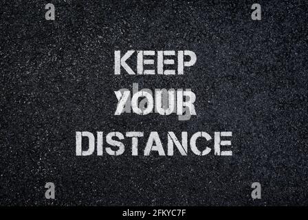 "Keep your distance" sign/text on dark, wet asphalt road, top view. Full frame textured background of social distancing sign on black asphalt. Stock Photo