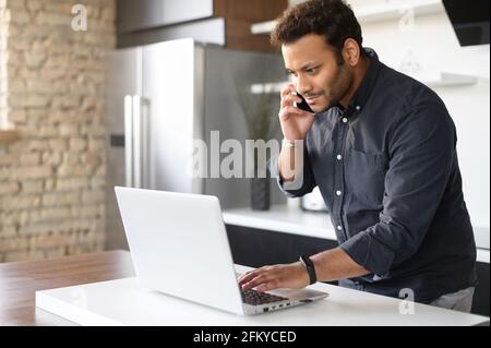 Busy mixed-race young man in smart casual shirt using laptop computer for remote work, a guy is talking on the phone and typing in same time standing in the kitchen at home, work on the distance Stock Photo