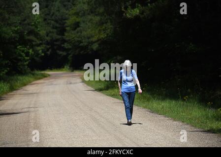 Elderly woman walks alone along a deserted country road.  Woods close in around her and road disappears into the distance. Stock Photo