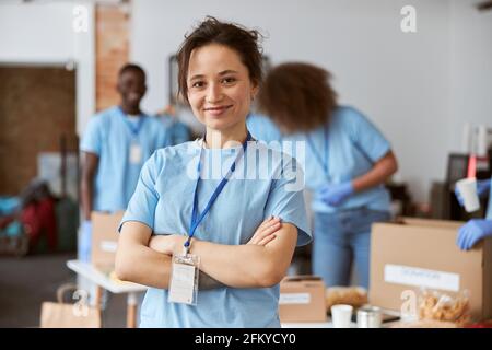 Portrait of happy young woman, volunteer in blue uniform smiling at camera while standing with arms crossed indoors. Team sorting, packing items in Stock Photo