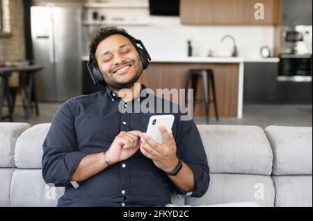 Cheerful handsome mixed-race guy wearing headset enjoys music sitting on the couch at home, indian man holding smartphone, listening favorite tracks with eyes closed, dreaming Stock Photo