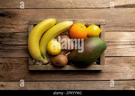 Fresh tropical fruits in a wooden delivery box on a wooden background. Orange, banana, mango, kiwi and lemon top view flat lay with copy space for som Stock Photo