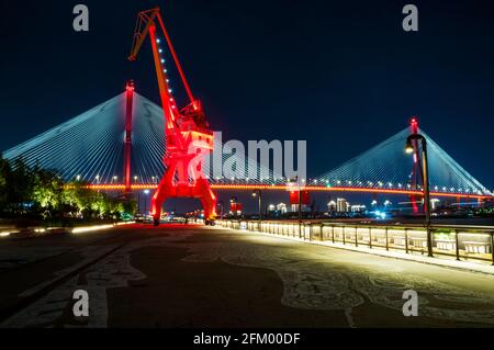 An old shipping crane on the Huangpu River waterfront at night with the Yangpu Bridge behind in Yangpu District, Shanghai, China. Stock Photo