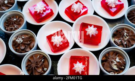 Red jelly dessert with whipped cream served on white plate and cupcakes with chocolate mousse in cup. Square red jelly plate on table at restaurant Stock Photo