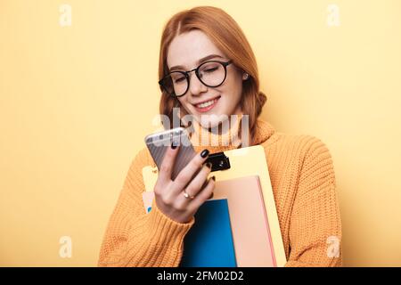 Freckles ginger student is smiling on a yellow wall chatting on mobile and holding folders Stock Photo