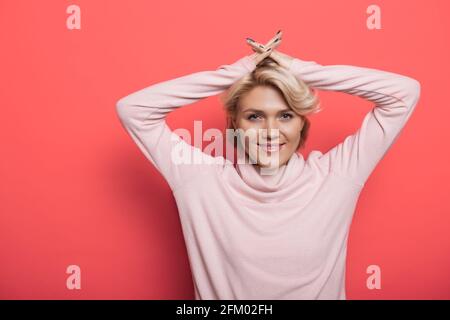 Charming blonde woman on a red studio wall is smiling at camera holding hands on head Stock Photo