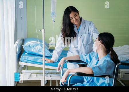 Smiling young Asian female doctor doing routine checkup and talking and comforting depressed patient sitting on wheelchair in hospital ward Stock Photo