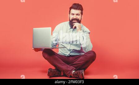 Smiling handsome bearded man worker laptop. Happy young man sitting on the floor with and using laptop computer on red background Stock Photo