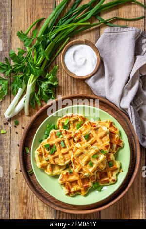 Not sweet waffles with potatoes, peas, bell peppers, cheese are served with sour cream and herbs on a plate on a rustic table. Top view flat lay. Stock Photo