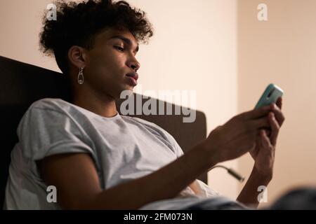 Social networks. Portrait of young guy with piercing using smartphone in his bed at night before going to sleep Stock Photo