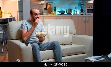 Sport football fan watching game late at night while enjoying popcorn, cheering for favourite football team. Championship entertainment match alone television, soccer fan looking at competition Stock Photo