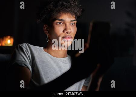 Portrait of handsome young guy using smartphone at night, sitting in dark room at home Stock Photo