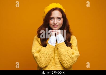 Happy young woman with long red hair in sweater and beret standing over yellow wall background Stock Photo