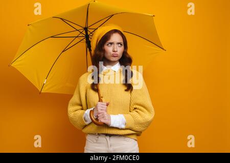 Happy young woman with long red hair wearing beret and sweater standing under umbrella over yellow background Stock Photo