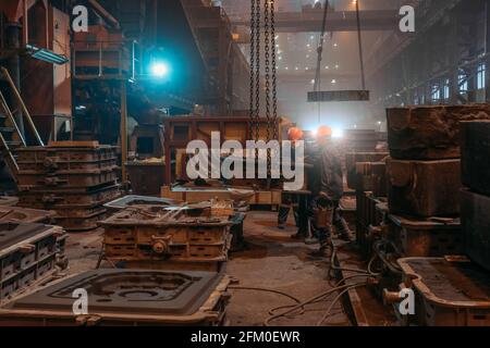 Workers in helmets and face masks works with mold on crane chains after metal casting in steel mill foundry. Stock Photo