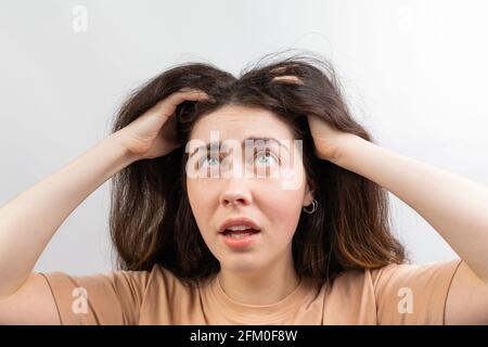 Dandruff, pediculosis and seborrhea. Portrait of a young Caucasian woman, a brunette in a beige t-shirt, who scratches her head with her hand in surpr Stock Photo