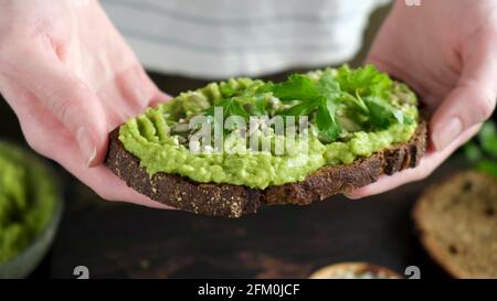 Female hands holding vegan rye bread avocado toast with seeds and parsley, closeup view Stock Photo