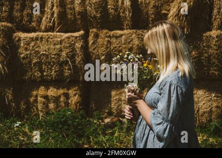 Young girl in rubber boots with flowers standing against the background of straw bales on country farm. Farming, Cottagecore, Farmcore, Countrycore Stock Photo