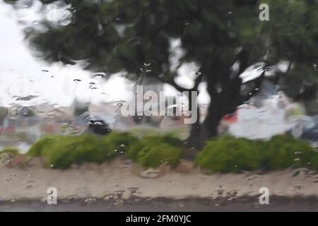 Rain drops hitting a wet windshield with a tree with green leaves and bushes during a rainy cloudy spring day Stock Photo