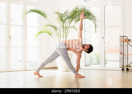 Low angle view of an athletic man doing a Parivrtta trikonasana or Triangle yoga pose stretching and strengthening his core muscles in a high key gym Stock Photo