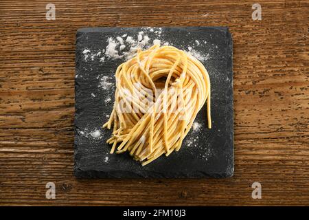 Top view of uncooked Italian handmade spaghetti alla chitarra egg pasta on black board sprinkled with flour placed on wooden table Stock Photo