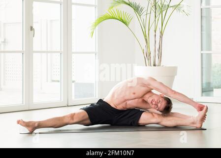 Young athletic man doing a Upavistha konasana pose or wide open forward stretch grasping his toes in a high key gym with copyspace in a health and fit Stock Photo