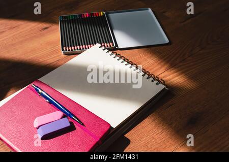 Image of a pink notebook, two erasers, a fill pencil, a sketchbook, and colored pencils framed by shadow Stock Photo