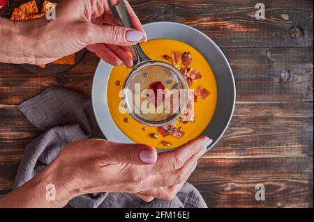 Woman's hands sprinkled with sieve paprika on traditional pumpkin puree cream soup with carrots, onions, ginger, garlic, coconut milk. Healthy, vegetarian food. Top view, selective focus Stock Photo