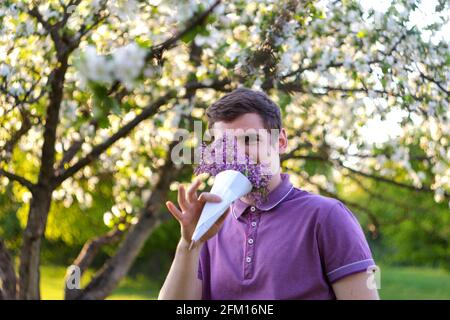 Defocus portrait of handsome caucasian man with cone flowers in lilac garden. Funny attractive young brunette guy smelling fresh flowers and looking a Stock Photo