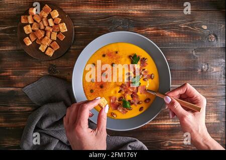 Woman's hands hold crouton and spoon with traditional pumpkin puree cream soup with carrots, onions, ginger, garlic, coconut milk. Added seeds, fried bacon, parsley. Healthy, vegetarian food. Top view Stock Photo