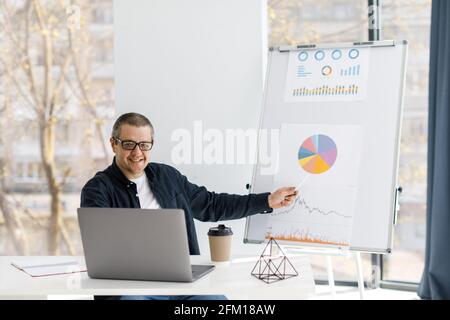 A man dressed in casual clothes works in the office, sits at a table near a laptop and holds an online conference. The man works in a remote place of Stock Photo