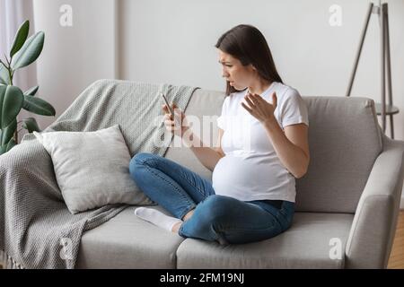 Stressed Young Pregnant Woman Looking At Smartphone Screen While Relaxing On Couch Stock Photo