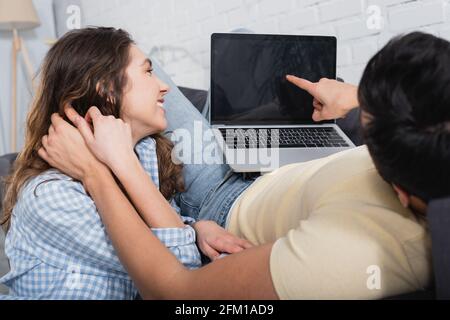 man pointing at laptop with blank screen near happy girlfriend Stock Photo