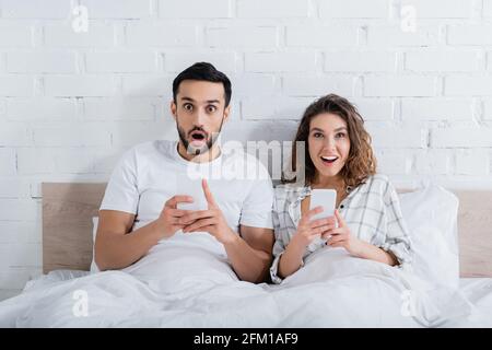 amazed interracial couple lying on bed and using smartphones Stock Photo