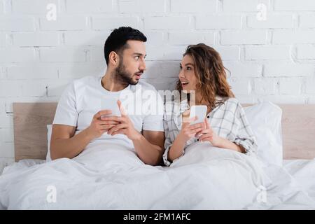 interracial couple looking at each other while lying on bed and using smartphones Stock Photo