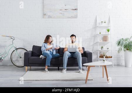 muslim freelancer using laptop near girlfriend with digital tablet on couch Stock Photo