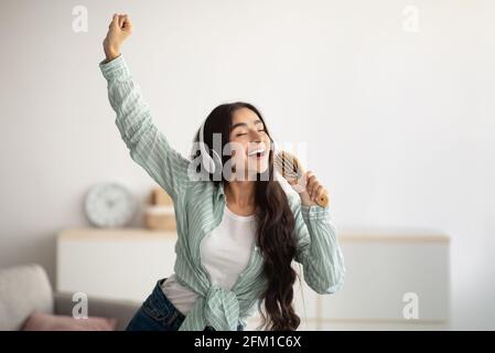 Happy Indian lady with headphones dancing to music and singing song, using hairbrush as mic at home Stock Photo