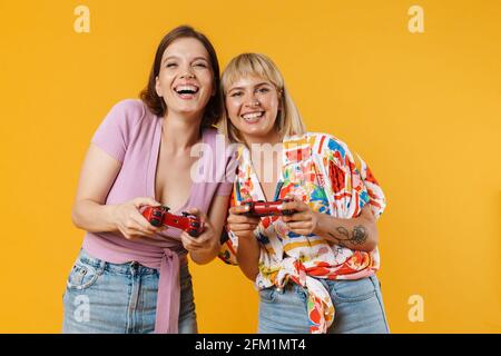 Portrait of two cheerful lovely girlfriends wearing summer clothes standing isolated over yellow background, holding joysticks Stock Photo