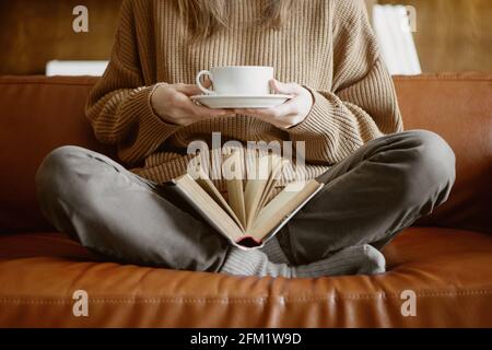 Cropped image of woman sitting on the couch holding cup of tea and reading a book. Stock Photo