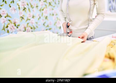 Closeup photo of dressmaker woman hands cutting yellow fabrics with blurred background. Professional instrument concept. Tailor woman cuts linen textile cloth in atelier boutique. High quality photo Stock Photo