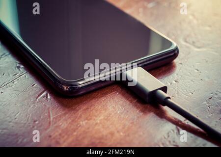 Black Smartphone With Connected Black USB Cable On A Brown Wooden Table Stock Photo