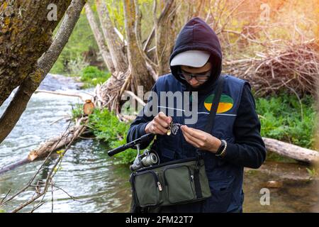 Young angler wearing a vest and warm clothes putting the hooks on the nylon and the rod to start fishing in the river where there are trees and fallen Stock Photo