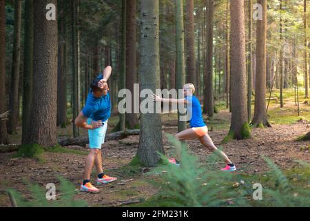 Couple doing warm-up exercises in the forest. A man doing shoulders and back stretching, a woman is leaning against the tree, doing calf exercises Stock Photo
