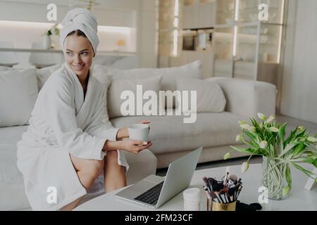 Young happy woman freelancer in bathrobe drinking tea and working remotely on laptop Stock Photo
