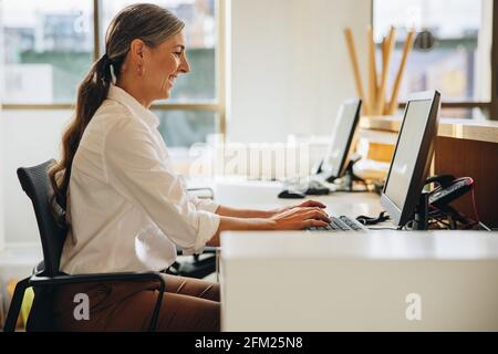 Side view of smiling woman sitting at desk and typing on computer in office. Mature woman smiling while working on computer. Stock Photo