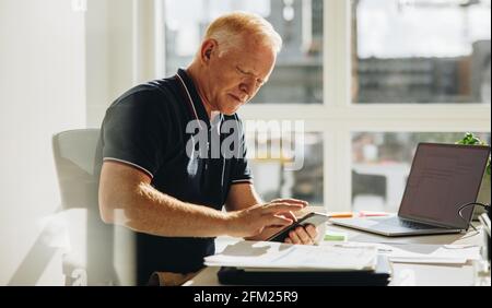 Senior businessman sitting at his desk and using mobile phone. Mature man reading text message on his cell phone at work. Stock Photo