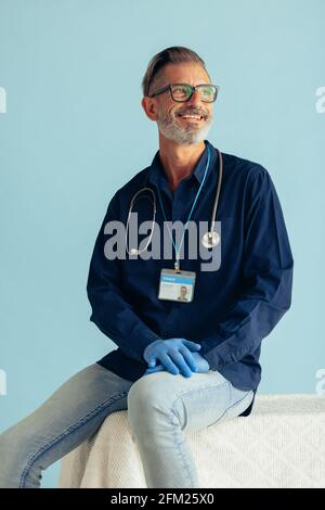 Mid adult doctor sitting on a table and looking away. Medical professional smiling on blue background. Stock Photo