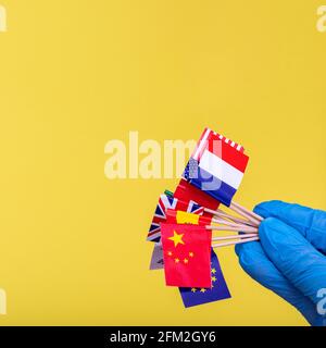 A hand in a blue protective glove holds copies of the flags of different countries on a bright yellow background, copy space, close-up, square frame. Stock Photo