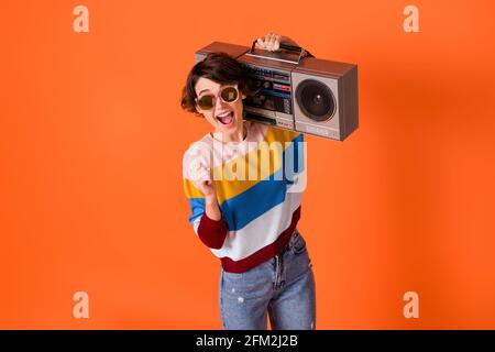 Photo of funky lucky young woman wear striped sweater eyewear rising fist holding boombox isolated orange color background Stock Photo
