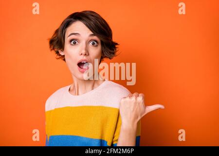 Photo of sweet impressed young woman wear striped sweater pointing thumb up empty space isolated orange color background Stock Photo
